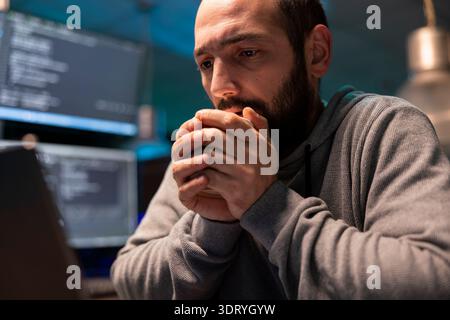 Tired software engineer warms his hands on hot coffee cup while thinking through late night debugging tasks. Concerned programmer stares at computer display, worried about system issues and deadlines. Stock Photo