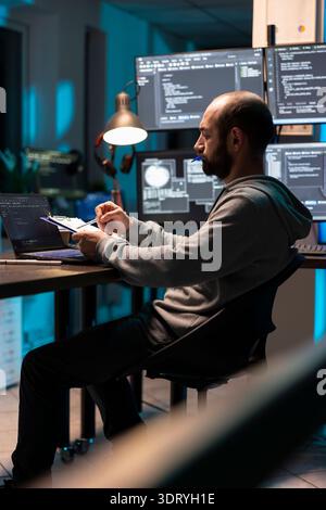 Adult male developer sits at workstation surrounded by digital monitors, holding pen cap while coding. Focused it specialist working on cybersecurity, network protection and cloud based systems. Stock Photo