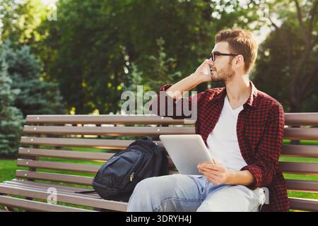 Guy Talking on Phone While Using Tablet in Park During Sunny Day Stock Photo
