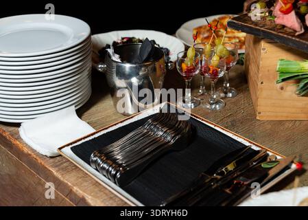 Rustic catering setup on a wooden table with a tall stack of white dinner plates, folded white napkins, and a tray of forks and knives on a black line Stock Photo