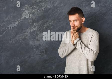 Young Man Stands With Hands Clasped Together in a Thoughtful Pose Against a Gray Wall Backdrop Stock Photo