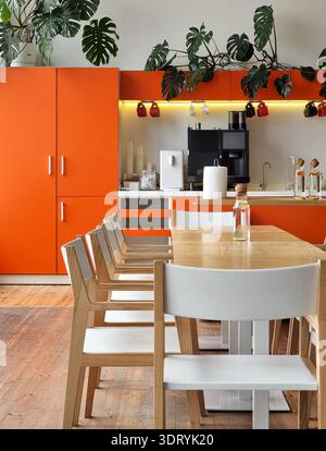 Modern coworking dining area with orange accents. Stylish kitchen featuring bright orange cabinetry, coffee machine, modern wooden furniture & plants. Stock Photo
