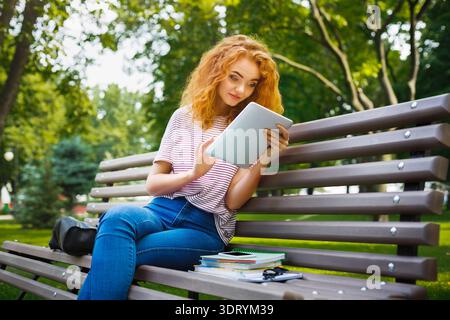Young Woman Reading on a Tablet While Sitting on a Bench in a City Park During Daytime Stock Photo