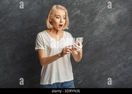 Woman Shows Surprise While Using Smartphone in Indoor Setting With Gray Background Stock Photo