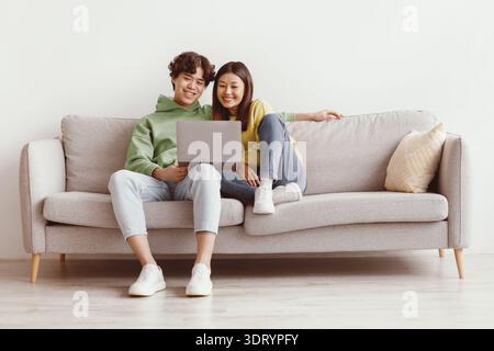 Happy young Asian couple using laptop, relaxing together on sofa at home, copy space Stock Photo
