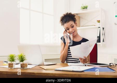 Woman Talks on Phone While Reviewing Papers at Office Desk During Daytime Stock Photo