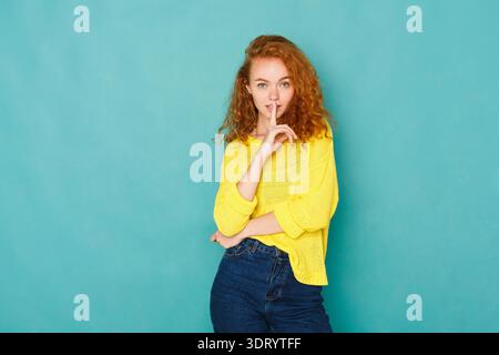 Young Woman in Yellow Sweater Making a Shushing Gesture in Front of a Blue Background Stock Photo