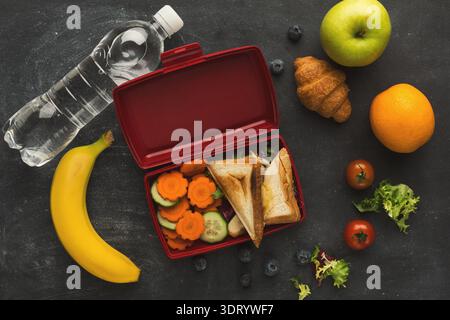Healthy Lunch Ideas With Fruits, Vegetables, and Sandwich in a Red Lunchbox on a Table Stock Photo