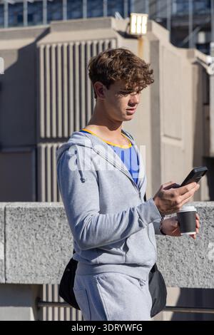 Man standing on rooftop in light gray hoodie and sweats, holding phone and paper cup Stock Photo