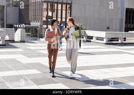 Two female students walking and talking on tiled campus plaza with folders backpacks headphones Stock Photo
