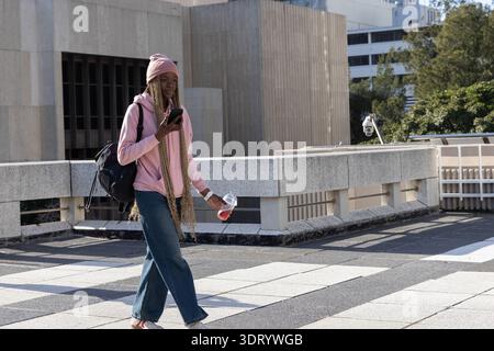 African American woman walking on tiled roof, wearing pink hoodie, checking smartphone, holding cup Stock Photo