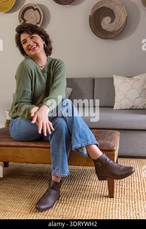 Woman sitting on low brown leather bench in cozy living room, smiling while wearing green sweater Stock Photo