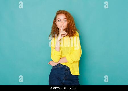 Young Woman With Curly Hair Holds Finger to Lips in Front of Blue Background Stock Photo