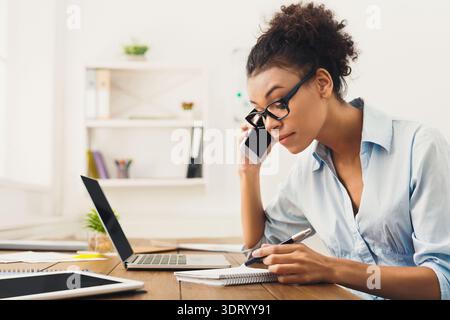 Woman Makes Phone Call While Taking Notes at Her Workspace in a Bright Office Stock Photo