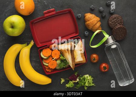 Healthy Lunch Collection With Fruits and Snacks in a Red Lunchbox Set on a Dark Surface Stock Photo