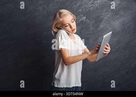 Young Woman Talks on the Phone While Using a Tablet in a Studio Setting With Gray Walls Stock Photo