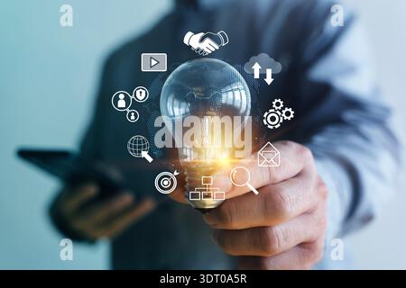 Close-up of businessman holding a glowing light bulb with digital icons for cloud, teamwork, security, analytics and communication, symbolizing innova Stock Photo
