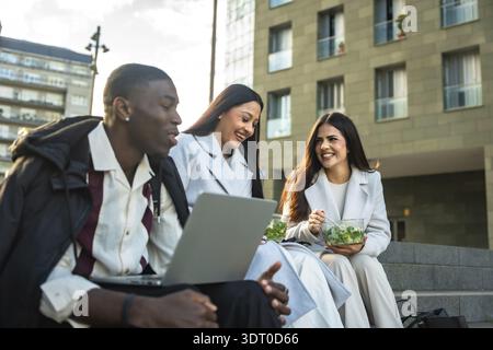 Three smiling diverse business people sitting on city stairs, discussing work and happy together during a successful outdoor lunch break with laptop a Stock Photo