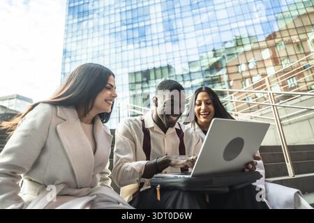 Group of diverse coworkers laughing while sharing content and collaborating on a laptop during an outdoor meeting in an urban setting, reflecting team Stock Photo