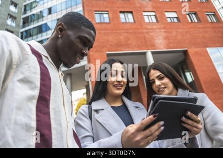 Startup business team including two women and a man collaborating and discussing work using a digital tablet while standing outside modern corporate b Stock Photo