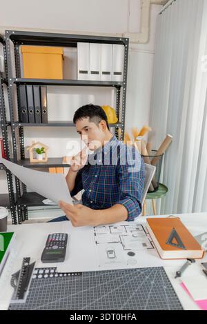 Young male architect working thoughtfully at office desk, analyzing house blueprints and construction plans, reviewing details carefully to ensure acc Stock Photo