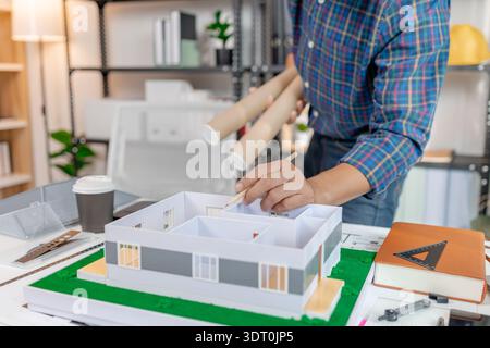 A young architect works with building plans and a house model in the office, reviewing blueprints, marking details, and analyzing design structures wi Stock Photo