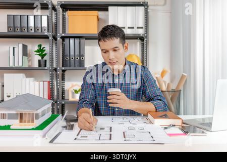 A young architect is working at his desk, reviewing blueprints, using a calculator, and checking a house model. He focuses on precision and detail whi Stock Photo