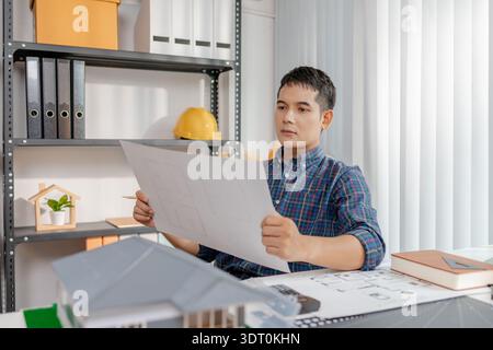 A young architect is working at his desk, reviewing blueprints, using a calculator, and checking a house model. He focuses on precision and detail whi Stock Photo