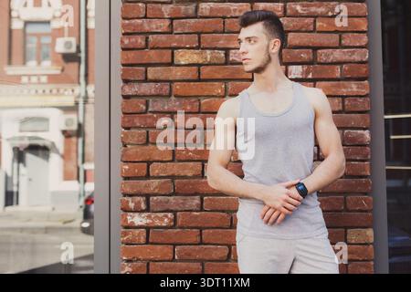 Young Man Stands by Brick Wall in Urban Area While Checking His Watch in the Afternoon Stock Photo
