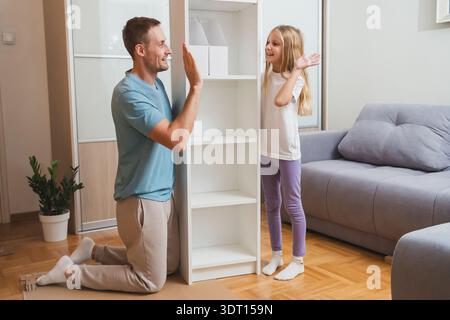 father and daughter give a cheerful high five while assembling a white shelf together on the floor, surrounded by tools and in a bright modern living Stock Photo