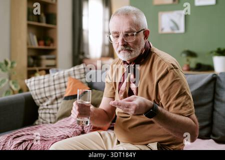 Senior Caucasian Man Sitting Indoors Holding Pills and Drinking Water Stock Photo