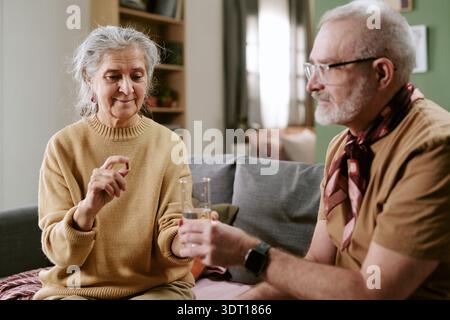 Senior Caucasian Woman Taking Pill While Senior Caucasian Man Offering Glass of Water Stock Photo
