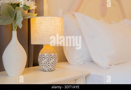 Stylish lamp and white flower pot on bedside table near bed indoors. Nightstand, lamp, and power outlet concept. Stock Photo