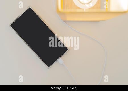 Top view of a smartphone while charging on the bedside table nearby lamp, connecting directily to the USB electrical socket built-in table. Copy space Stock Photo