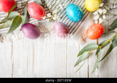 Bright pastel Easter eggs with eucalyptus leaves and white flowers on textured fabric over wooden background. Warm natural light and festive spring mo Stock Photo