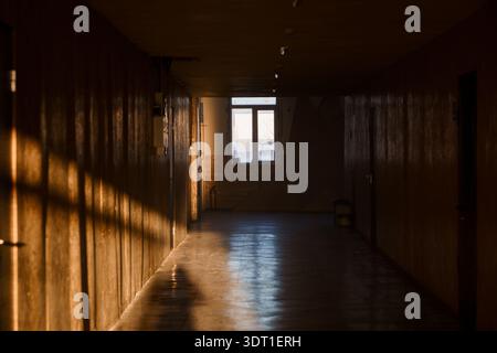 A dimly lit hallway with wooden walls and floor, illuminated by warm sunlight through a distant window, casting long shadows and creating a moody, atm Stock Photo