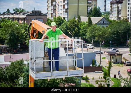 Portrait of smiling man working in elevated lift bucket of crane, wearing green cap and green shirt. Stock Photo