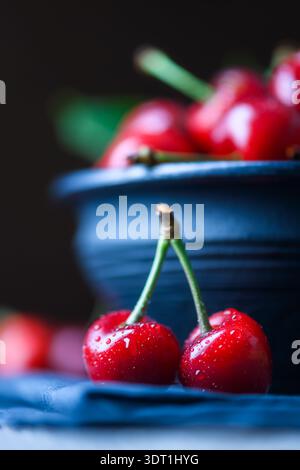 Two sweet cherries in front of dark bowl, selective focus composition. Fresh red summer fruit with stems, minimal food styling Stock Photo