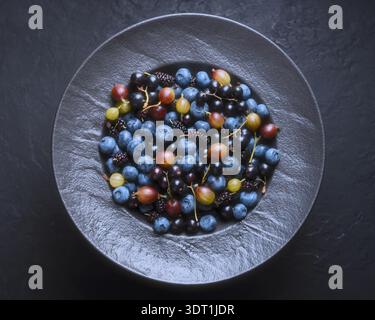 Mixed blue berries in grey plate on dark surface, overhead composition. Fresh blueberries, gooseberries and mulberries, clean modern food photography Stock Photo