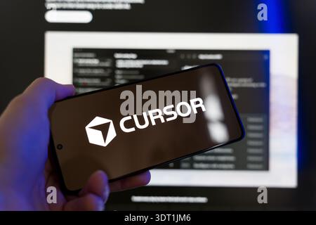 Hand holding smartphone displaying Cursor AI logo on screen with a blurred code editor in the background, artificial intelligence coding assistant and Stock Photo