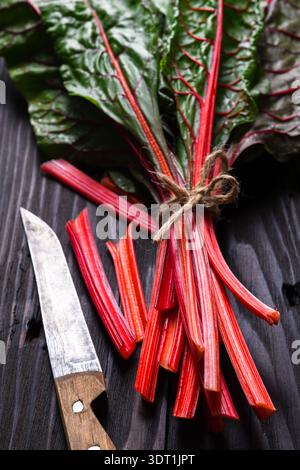 Fresh Swiss chard bunch with red stems tied with string on dark wooden surface. Organic leafy vegetable with knife, rustic food background Stock Photo