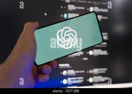 Hand holding smartphone displaying the OpenAI logo on screen with a blurred ChatGPT interface in the background, generative artificial intelligence an Stock Photo