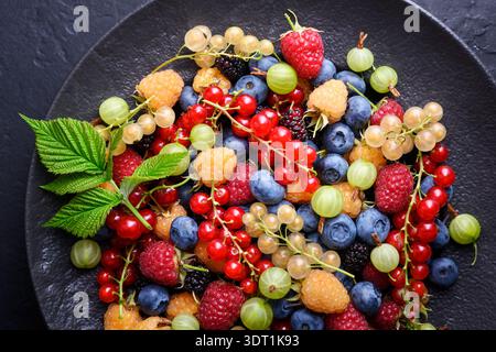 Colorful berry mix arranged in round plate on dark surface. Summer fruit assortment without cherries, vibrant natural food background Stock Photo