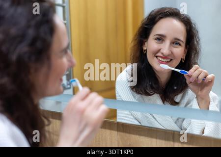 Smiling woman brushing teeth in bathroom mirror. Young woman in a bathrobe brushes her teeth and smiles at her reflection in a modern bathroom Stock Photo