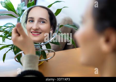 Smiling woman looking in mirror at home. Happy young woman checks her face in a small tabletop mirror, natural makeup and bright daylight with green Stock Photo