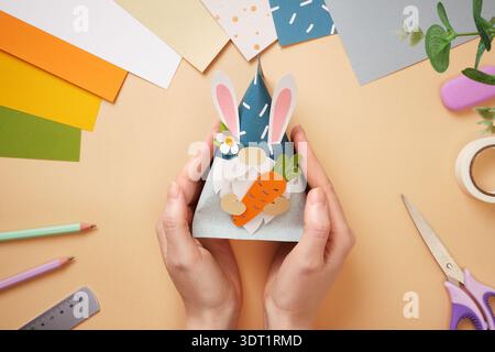 Hands holding finished paper craft of a gnome with a carrot. Tape, scissors, pencil on beige background. DIY handmade project for children. Stock Photo