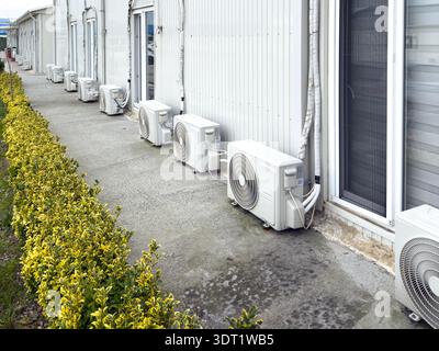 A series of outdoor air conditioning units installed on a building's exterior wall with a neatly trimmed hedge lining the paved walkway, showcasing ef Stock Photo