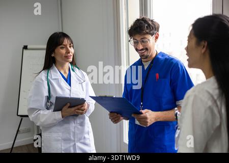A diverse medical team of two doctors and a nurse engaged in a discussion within a hospital. The professionals are wearing scrubs and lab coats, holdi Stock Photo