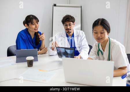 A collaborative medical team discusses patient care, featuring a nurse in white and two doctors, gathered around a table with medical documents and eq Stock Photo