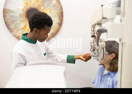 An optometrist in a white coat conducts a detailed eye examination using specialized equipment in a modern clinic setting with a large eye model in th Stock Photo